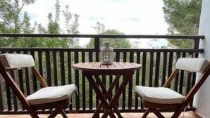 A balcony with two wooden chairs and a small table, a glass lantern, and a view of trees and a cloudy sky.