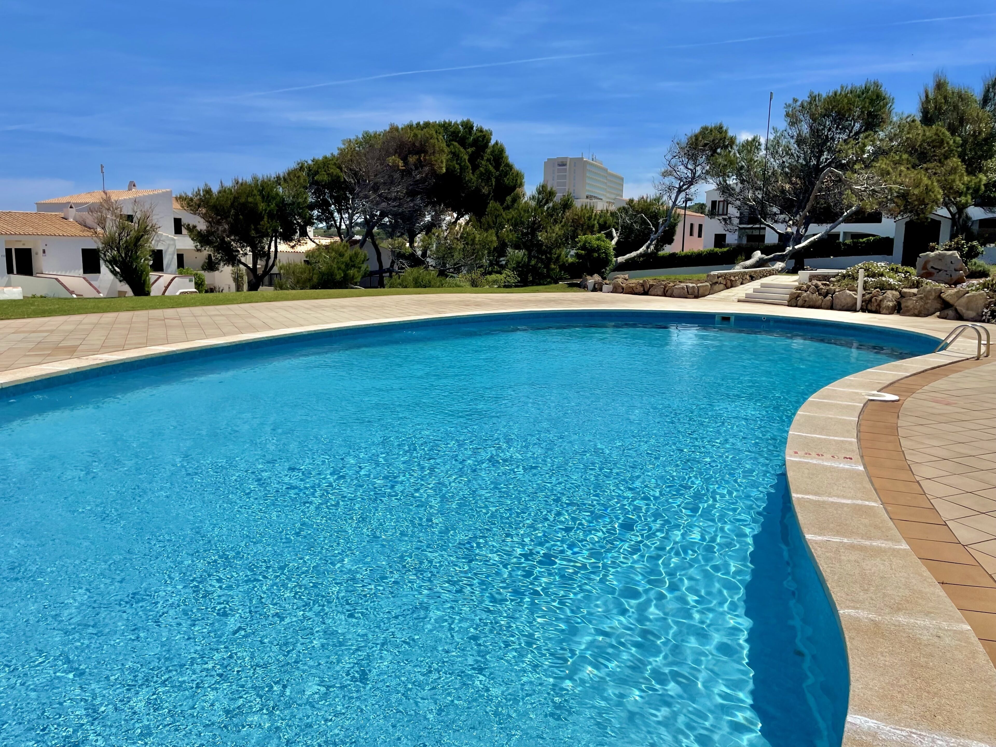 A clear blue swimming pool with a tiled deck, surrounded by greenery and buildings.