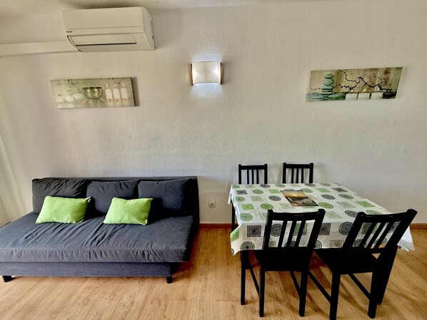 A room with a sofa, a dining table with chairs, an air conditioner, and wall art.