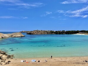 Ein Küstenstrand mit klarem türkisblauem Wasser, Felsformationen und einer entfernten, felsigen Insel mit Gebäuden.