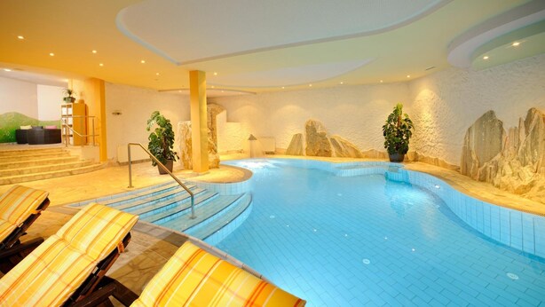 Una piscina interior con un fondo de pared de piedra, una escalera que conduce a una zona de estar con tumbonas y plantas en macetas.