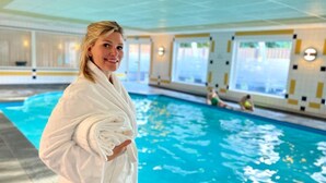 A person in a white bathrobe standing by an indoor swimming pool with clear blue water. - De Stobbe hotel & suites (Ruinen)
