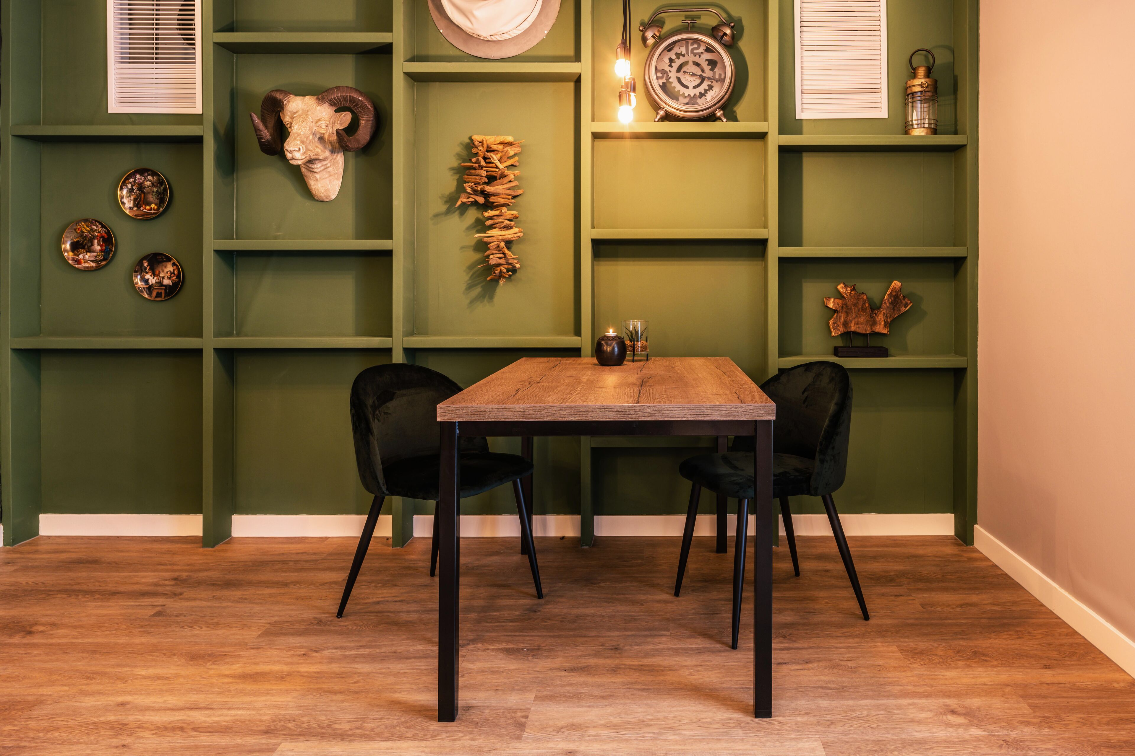 Un espace repas avec une table en bois et des chaises noires, entouré de murs verts ornés d’objets décoratifs.