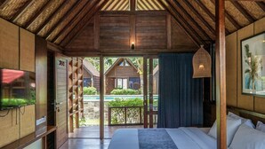 A room with a wooden ceiling, a bed with white and blue bedding, a TV on the left, and a view of the outside.