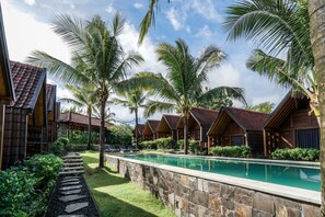 A pool area with a stone pathway, palm trees, and wooden cabins with red roofs. - Bingin Lodge Uluwatu (Pecatu)