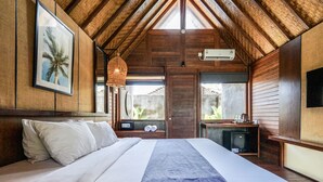 A bedroom with a large bed, wooden ceiling beams, a hanging light fixture, a small table with a radio, and a window with a view of the outside.