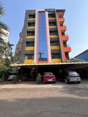 A multi-story hotel with a central glass elevator shaft, a parking area with several cars, and a small entrance canopy.