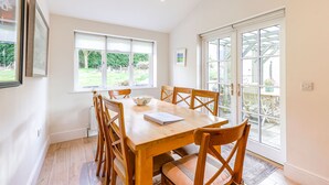 A dining room with a wooden table and chairs, large windows, and a view of the garden.