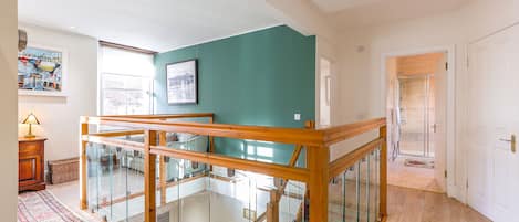 A spacious hallway with a wooden staircase leading to an upper floor, a glass railing, and a view of a door leading to a bathroom.