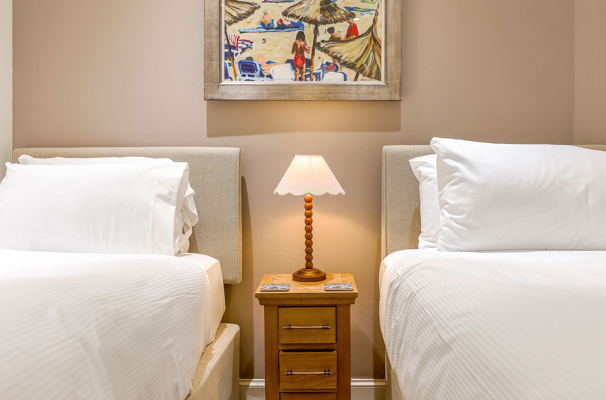 Een hotelkamer met twee bedden, een nachtkastje met een lamp en een schilderij aan de muur.
