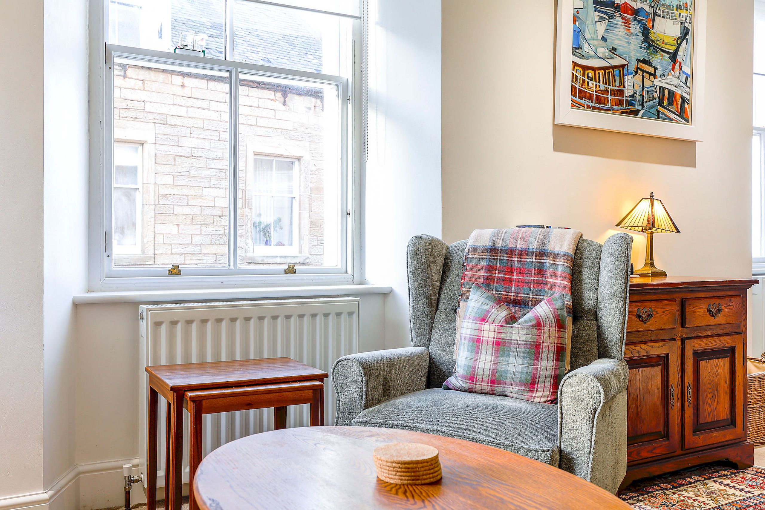 Une pièce chaleureuse avec une grande fenêtre, un fauteuil recouvert d’un plaid, une table en bois et une lampe.