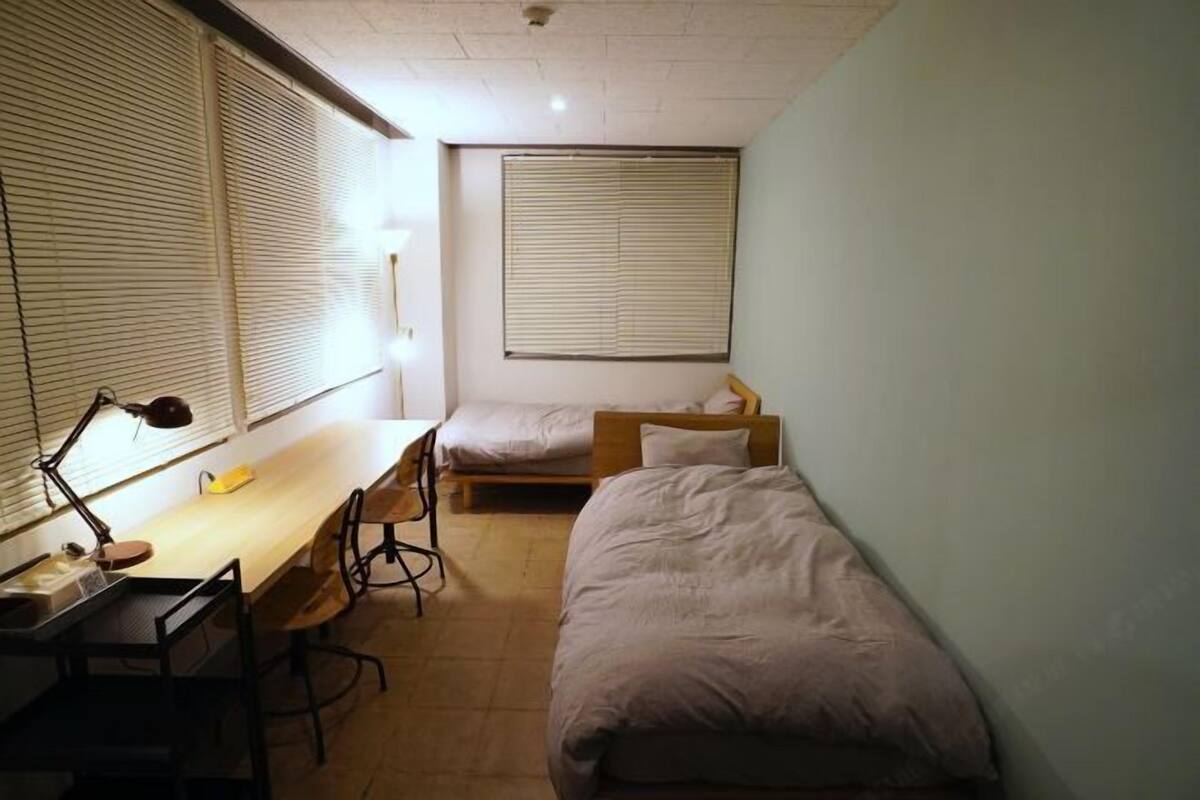 Habitación tipo dormitorio con escritorio, dos camas individuales, una ventana con persianas y una lámpara en el techo.