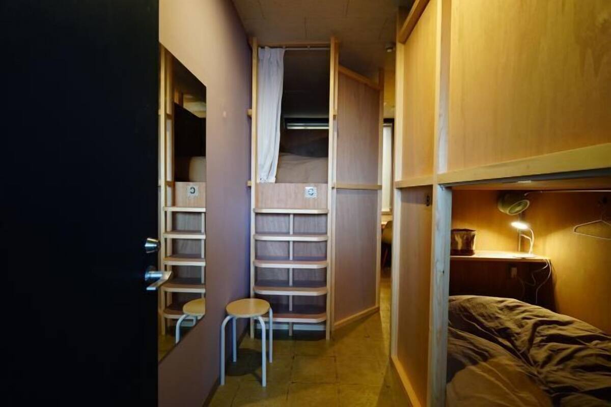 Una habitación pequeña con una cama litera, un taburete, un escritorio con lámpara y estanterías.