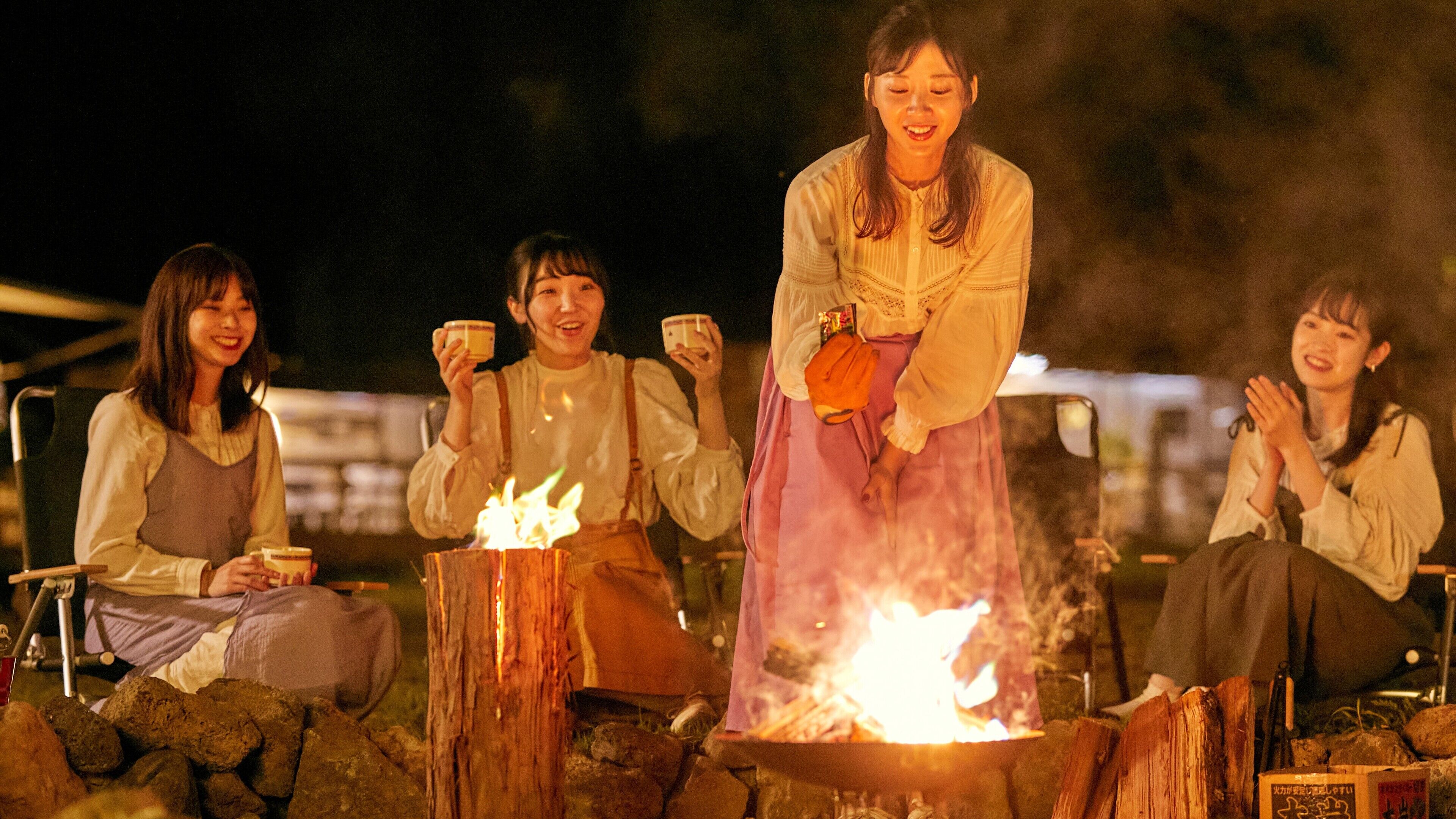 Four individuals seated around a campfire, with one person standing. They are holding cups, suggesting a social gathering.