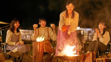 Four individuals seated around a campfire, with one person standing. They are holding cups, suggesting a social gathering.