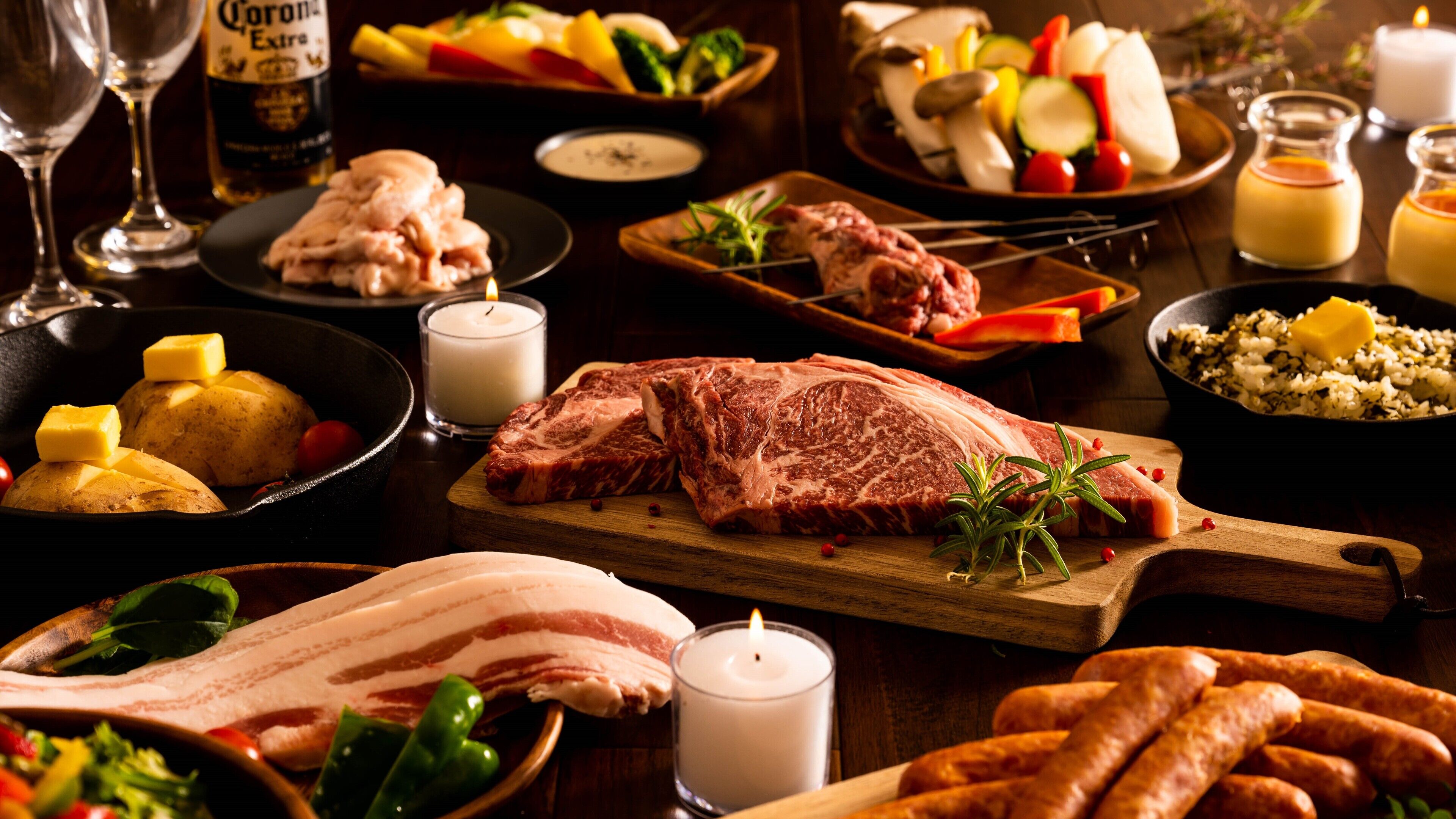 A variety of meats, vegetables, and condiments on a wooden cutting board, with a candle and glasses in the background.