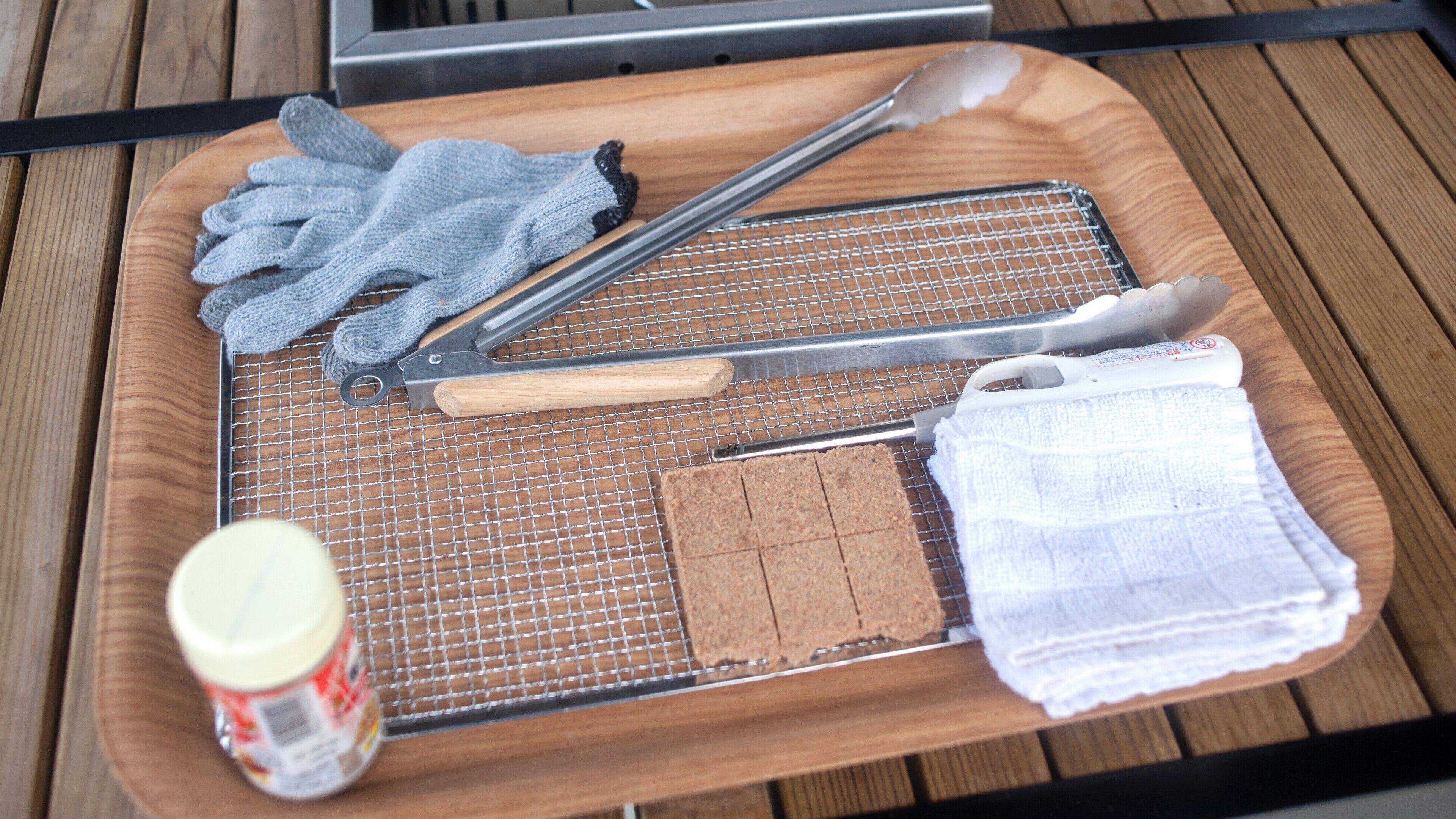 A tray with kitchen utensils, a cutting board, a jar of jam, and a white cloth on a wooden surface.
