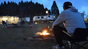 Un camping con una hoguera, tiendas de campaña y una caravana al fondo.