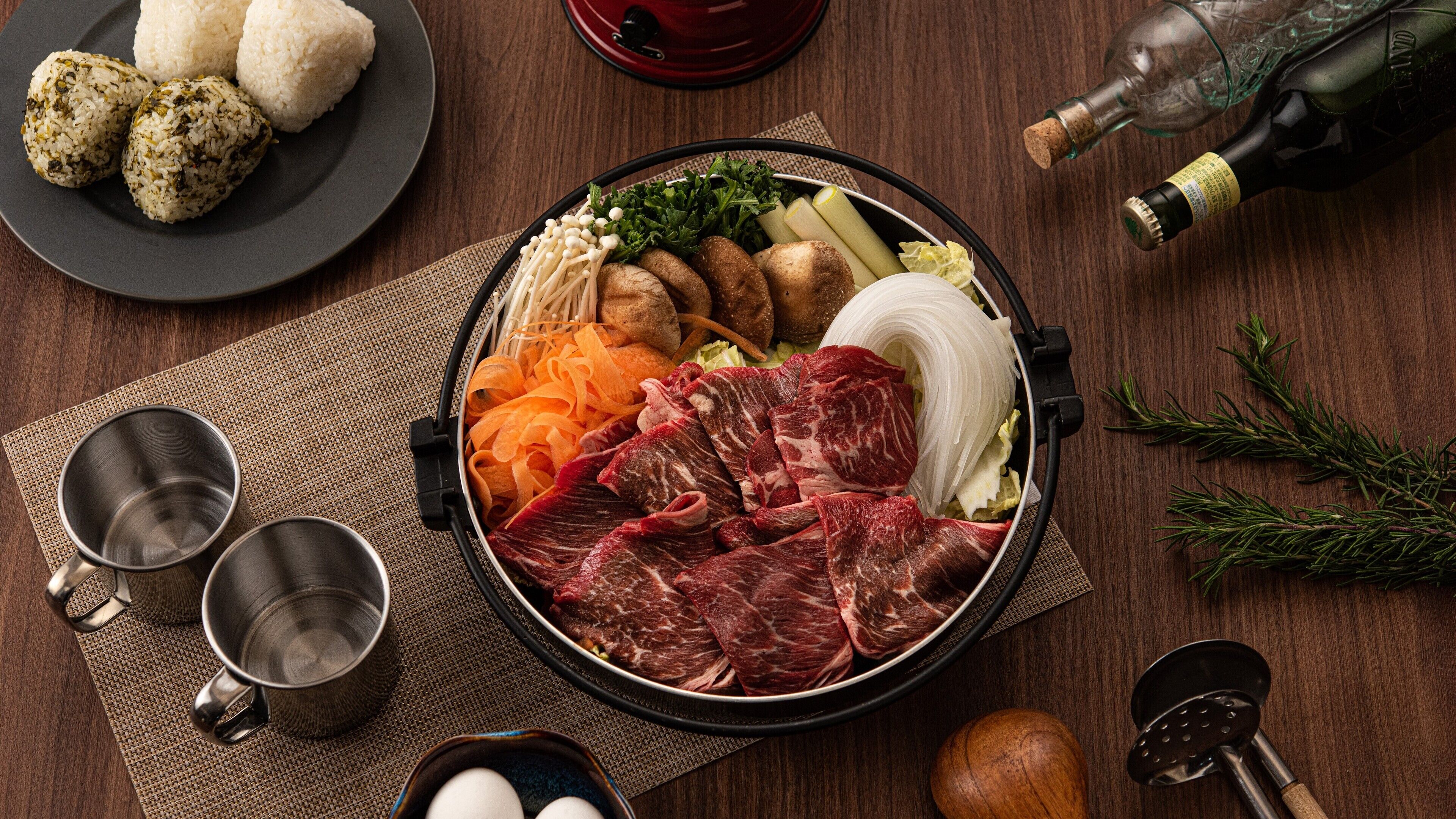 Een hete pot met diverse ingrediënten, een fles wijn en twee metalen bekers op een houten tafel.