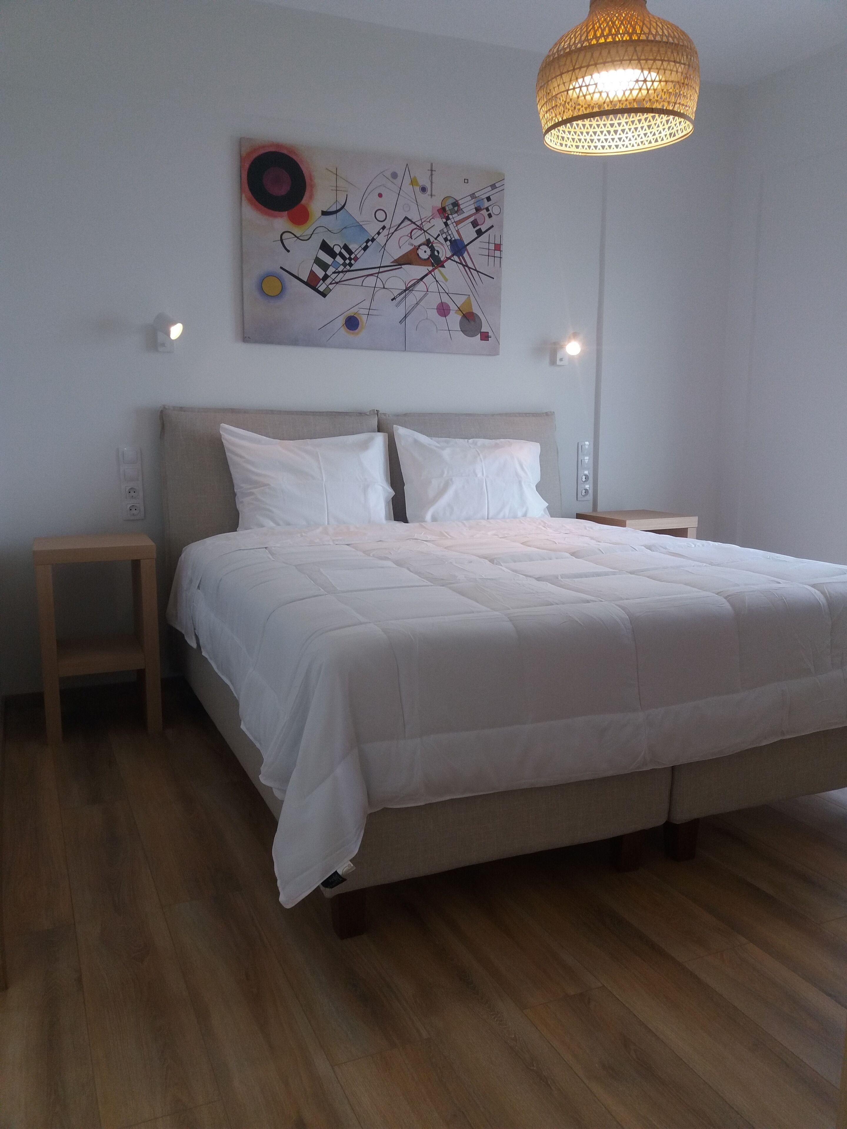 A bedroom with a bed, two bedside tables, a hanging light fixture, and a wall-mounted abstract painting.