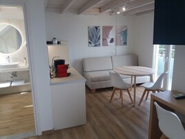 Una cocina moderna con un lavamanos, una encimera y una estufa. Hay un sofá y una mesa de comedor con sillas. La habitación tiene piso de madera y un ventanal grande.