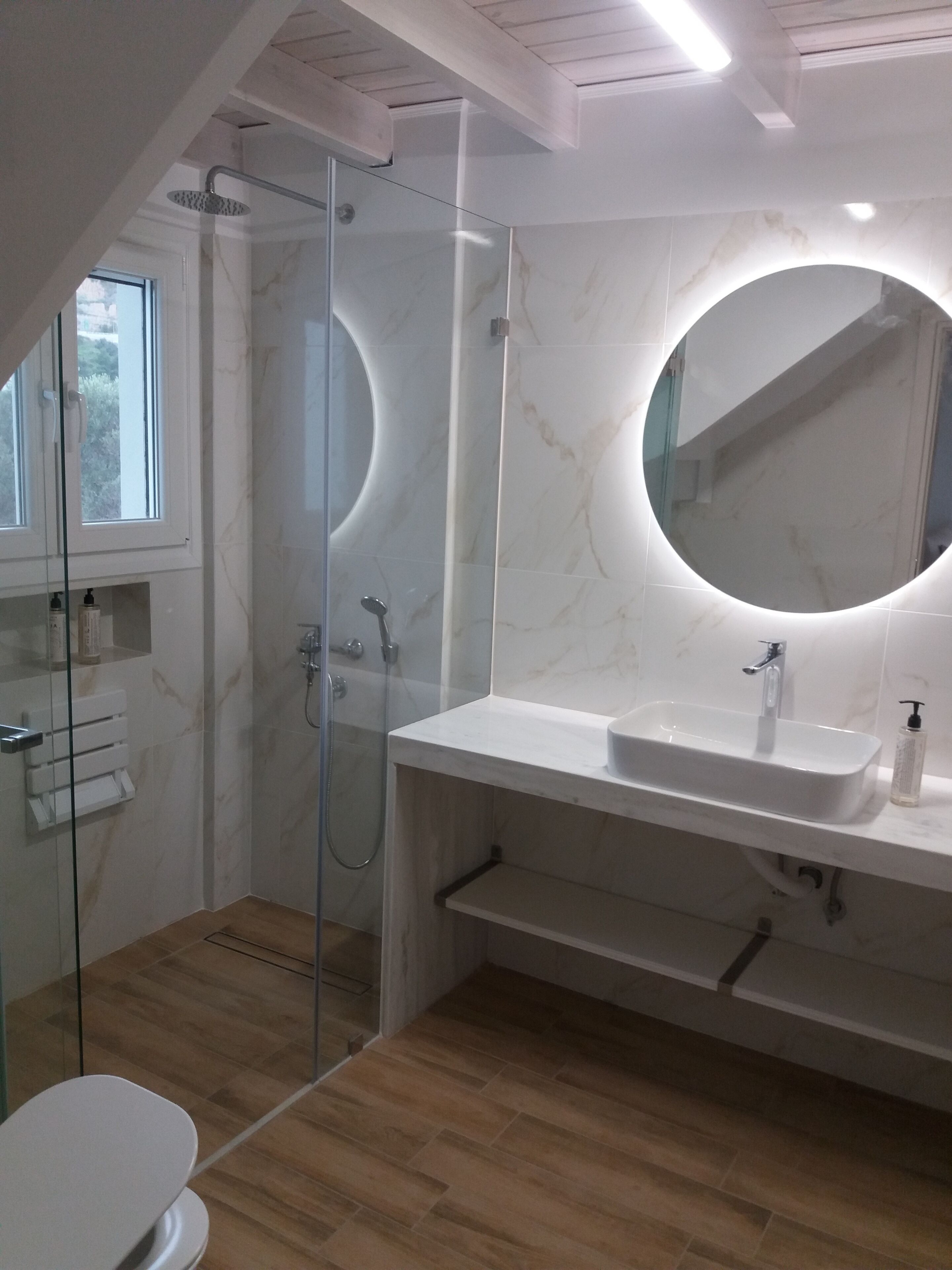 Un baño moderno con cabina de ducha de vidrio, un lavabo blanco con espejo redondo y suelo de madera.