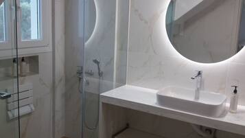 Un baño moderno con cabina de ducha de vidrio, un lavabo blanco con espejo redondo y suelo de madera.