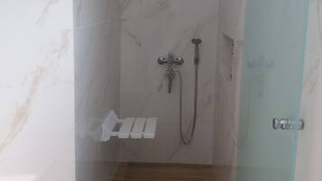 A small, enclosed shower area with a glass door, a handheld showerhead, a wall-mounted faucet, and a shelf with toiletries.