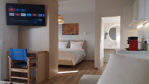 A hotel room with a bed, a desk with a chair, a TV mounted on the wall, and a small kitchenette area. - Stavento beach front suites (Aigialeia)