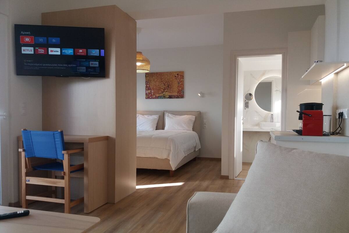 A hotel room with a bed, a desk with a chair, a TV mounted on the wall, and a small kitchenette area.