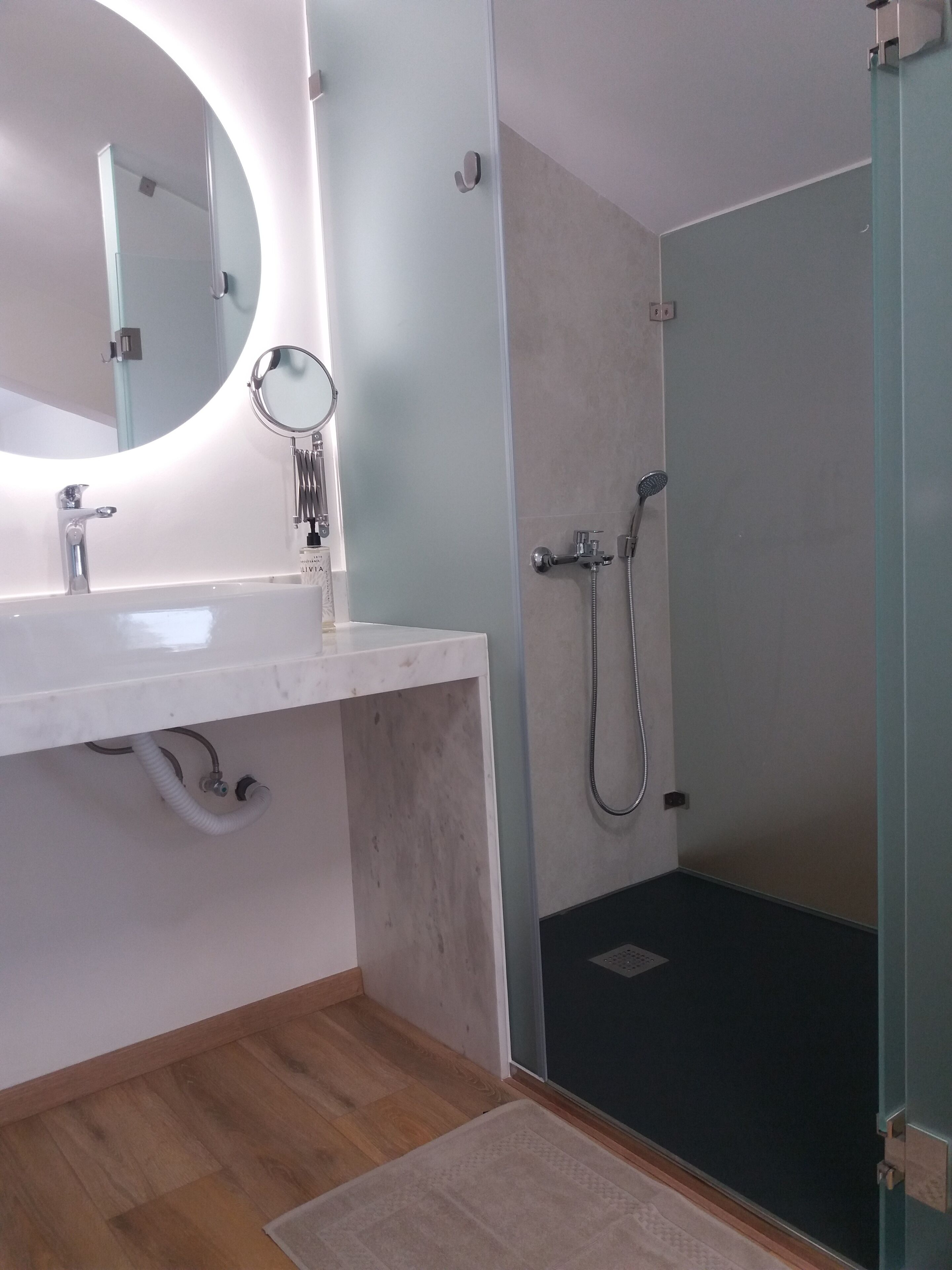 Un baño moderno con un lavamanos blanco, un espejo redondo, un cabezal de ducha manual y una ducha con mampara de vidrio.