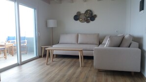 A modern living room with a sofa, a coffee table, and a lamp. There is a large window and a door leading to an outdoor area. - Stavento beach front suites (Aigialeia)