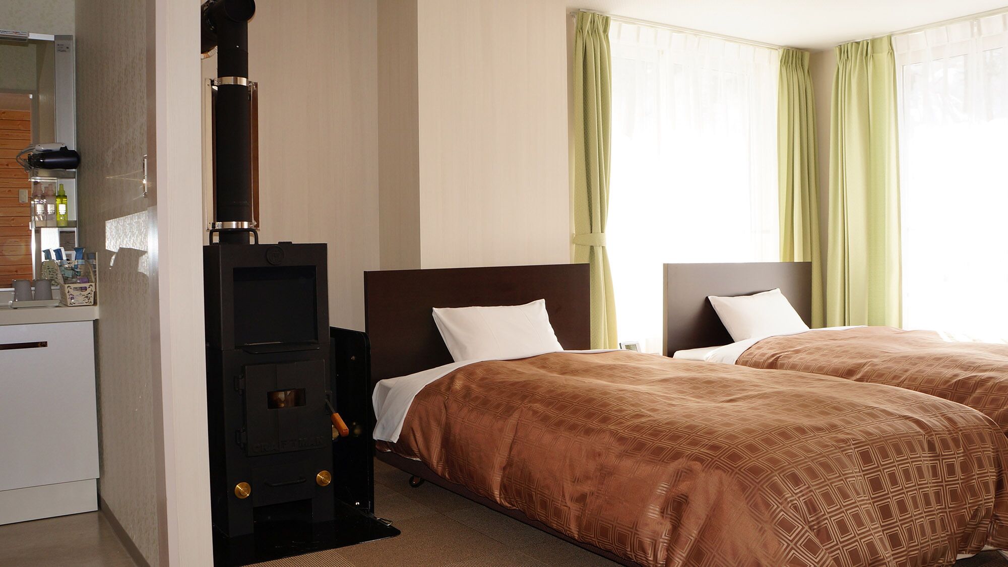 Habitación de hotel con chimenea de leña, dos camas con ropa de cama marrón estampada, una kitchenette pequeña y cortinas verdes.