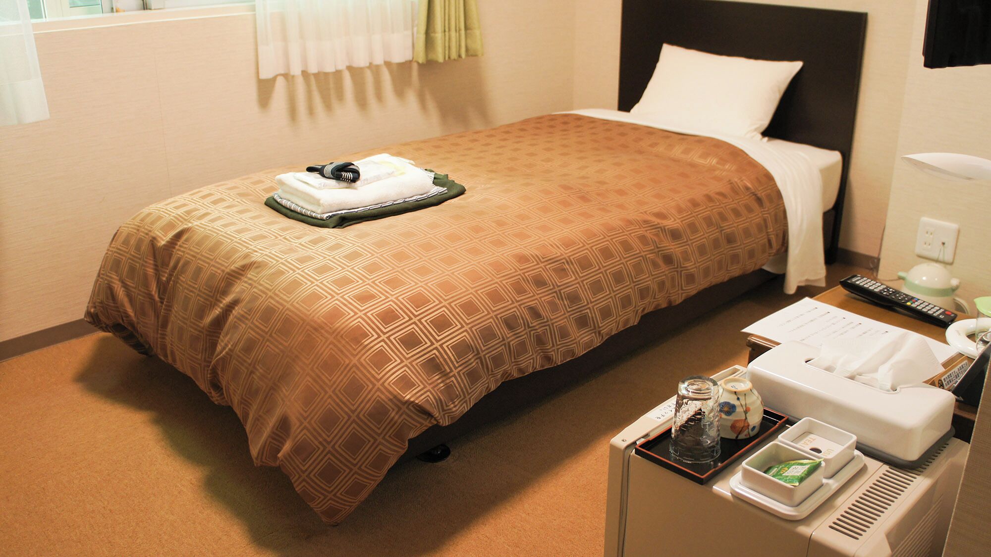 A hotel room with a bed, a bedside table with a remote control, a phone, and a tray with toiletries.