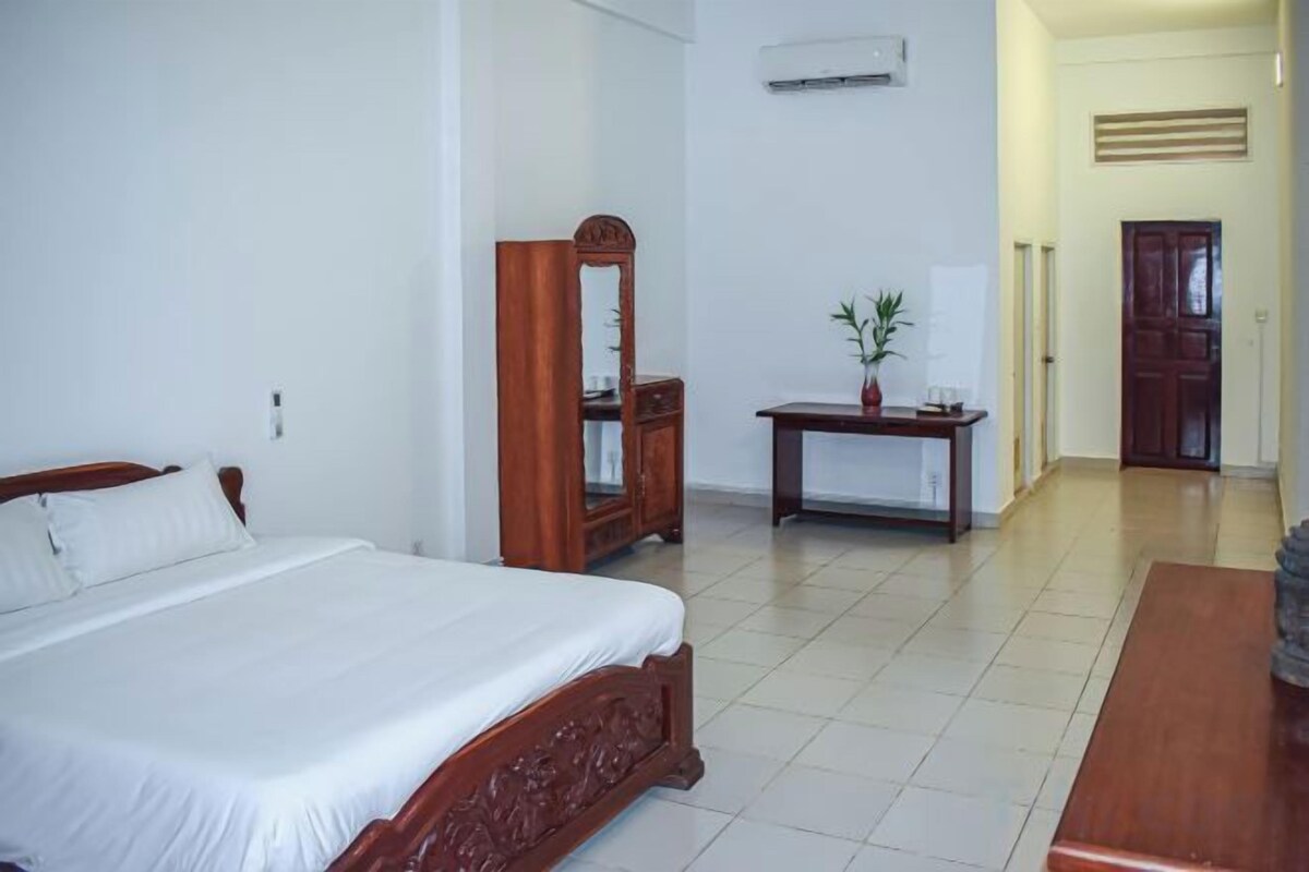 A hotel room with a bed, a wooden dresser, a small table with a plant, and a door leading to another room.
