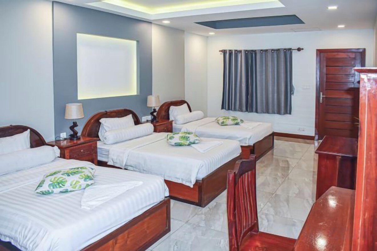 A hotel room with three beds, a wooden bench, a desk, and a large window with curtains.