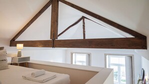 Una camera da letto con soffitto spiovente, travi in legno, un letto con lenzuola bianche, una lampada da comodino e una finestra.