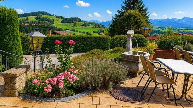 A terrace with outdoor seating, a stone fountain, and a view of a green valley and mountains. - Hotel JMS Holiday Allgäu (Oberreute)