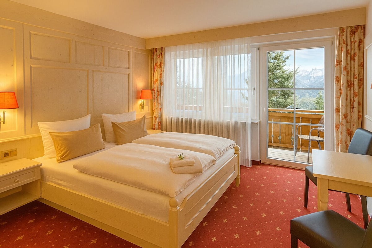 A hotel room with a double bed, two bedside tables with lamps, a small table with a chair, and a balcony with a view of mountains.