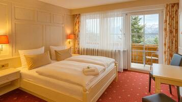 A hotel room with a double bed, two bedside tables with lamps, a small table with a chair, and a balcony with a view of mountains.