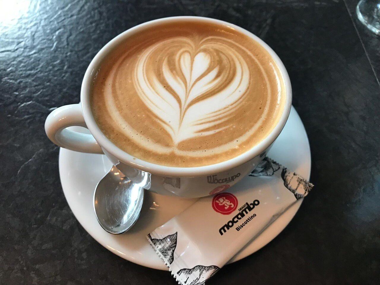 A cup of coffee with latte art on a saucer, a spoon, and a packet of biscotti.