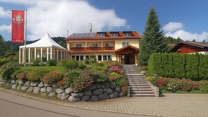 A two-story building with a stone wall and a garden with colorful flowers. - Hotel JMS Holiday Allgäu (Oberreute)