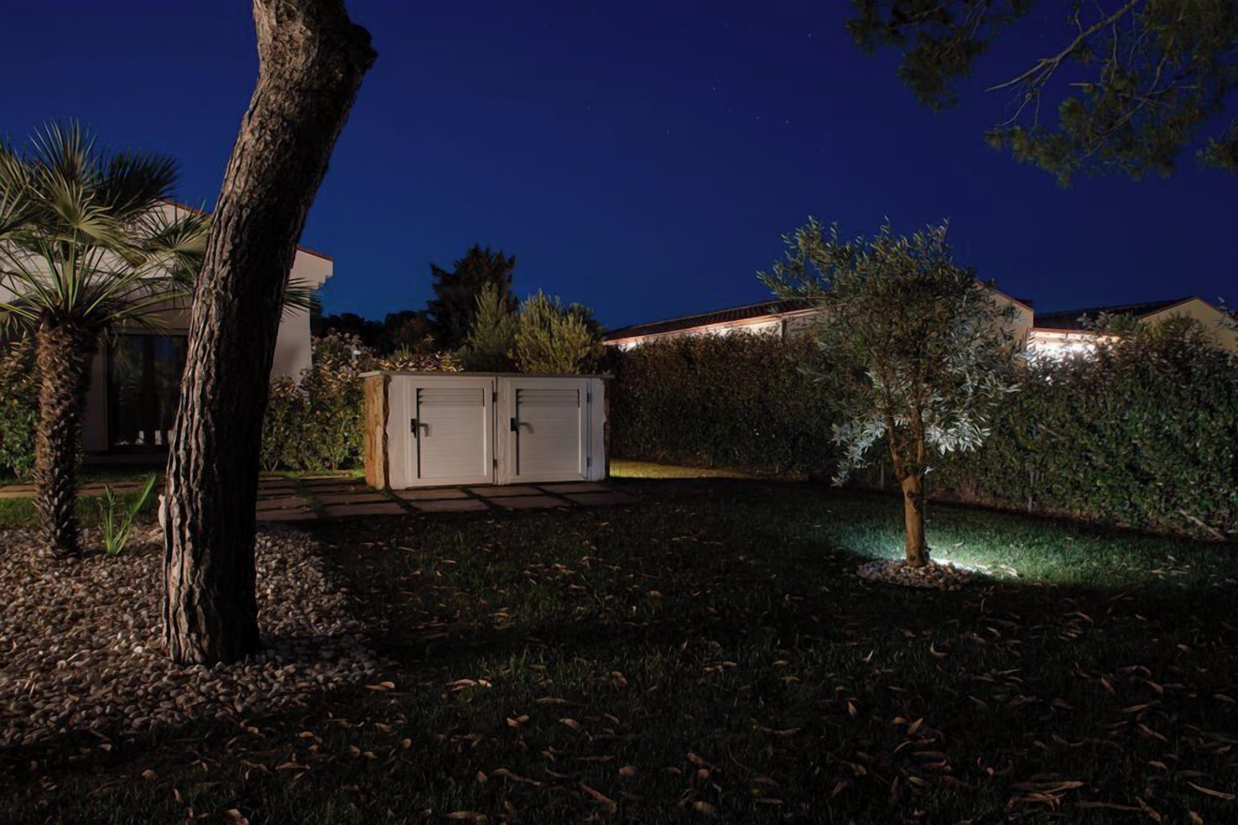 Une maison d'habitation la nuit, avec une pelouse bien entretenue, un abri de jardin et un arbre.
