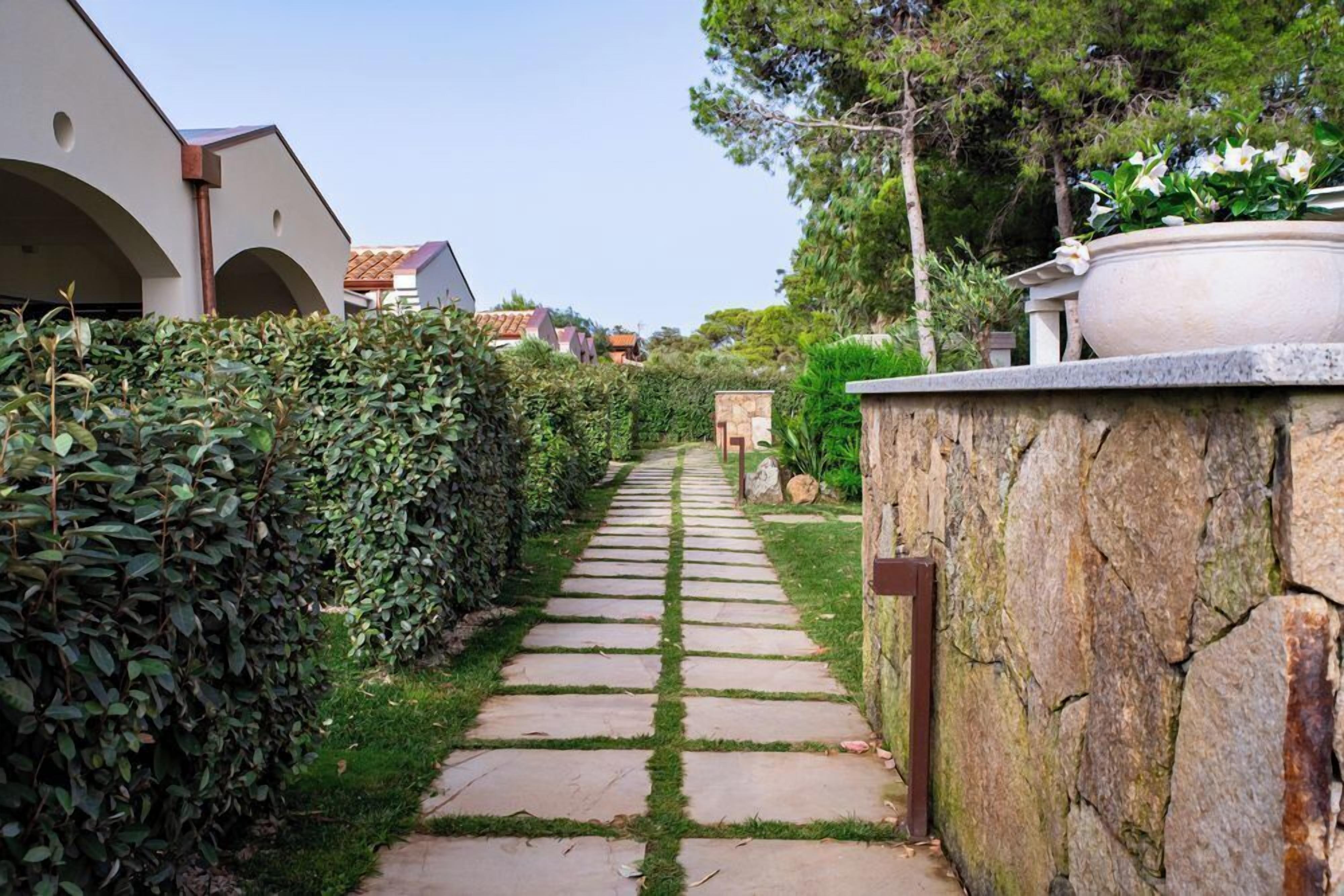 A stone pathway leading through a garden with a stone wall on one side and a hedge on the other.