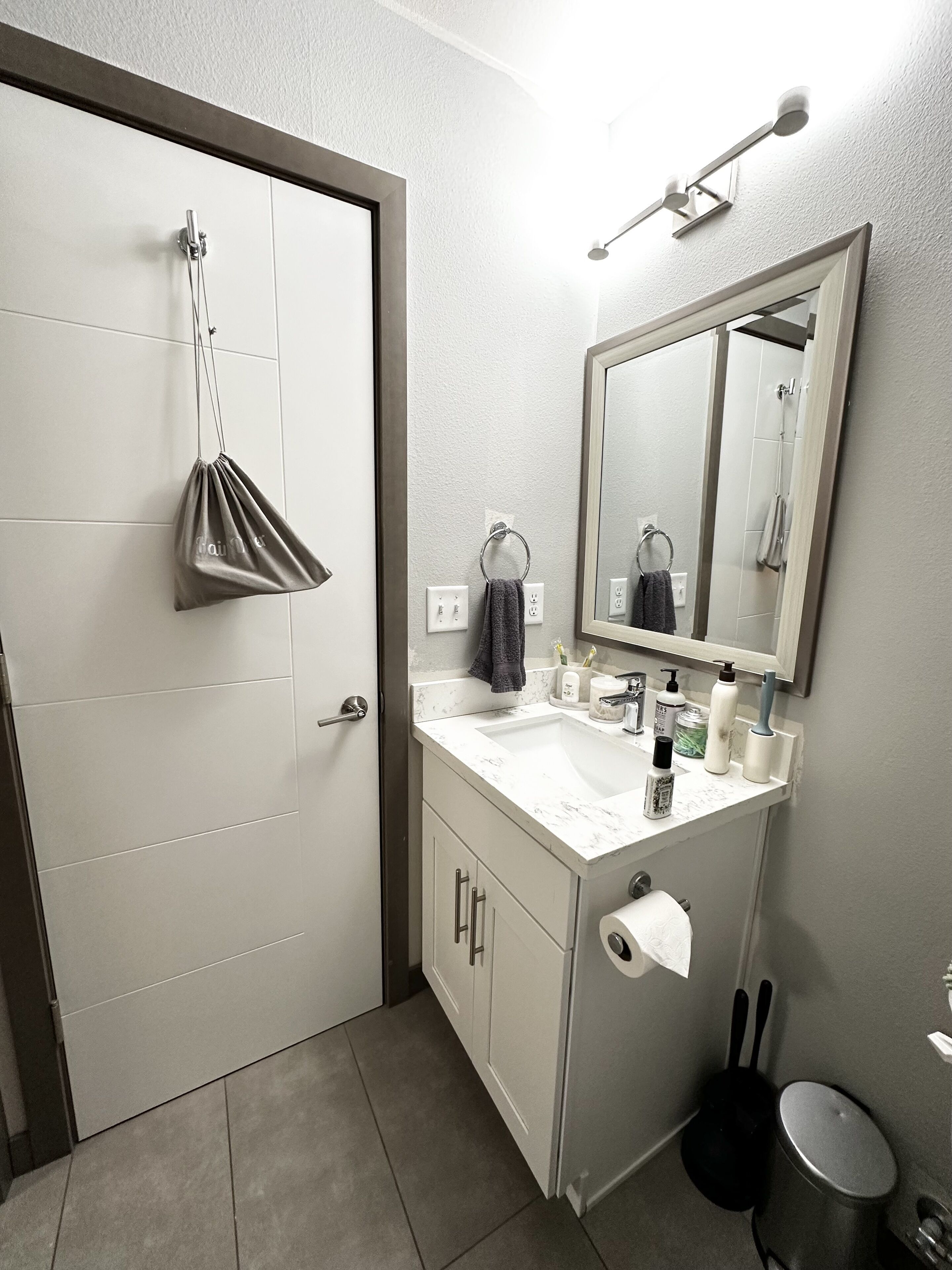 A bathroom with a white door, a sink with a mirror above it, a towel hanging on a ring, and a trash can.