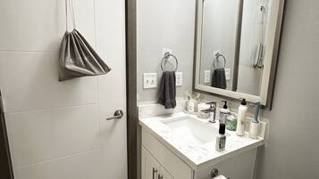 A bathroom with a white door, a sink with a mirror above it, a towel hanging on a ring, and a trash can.