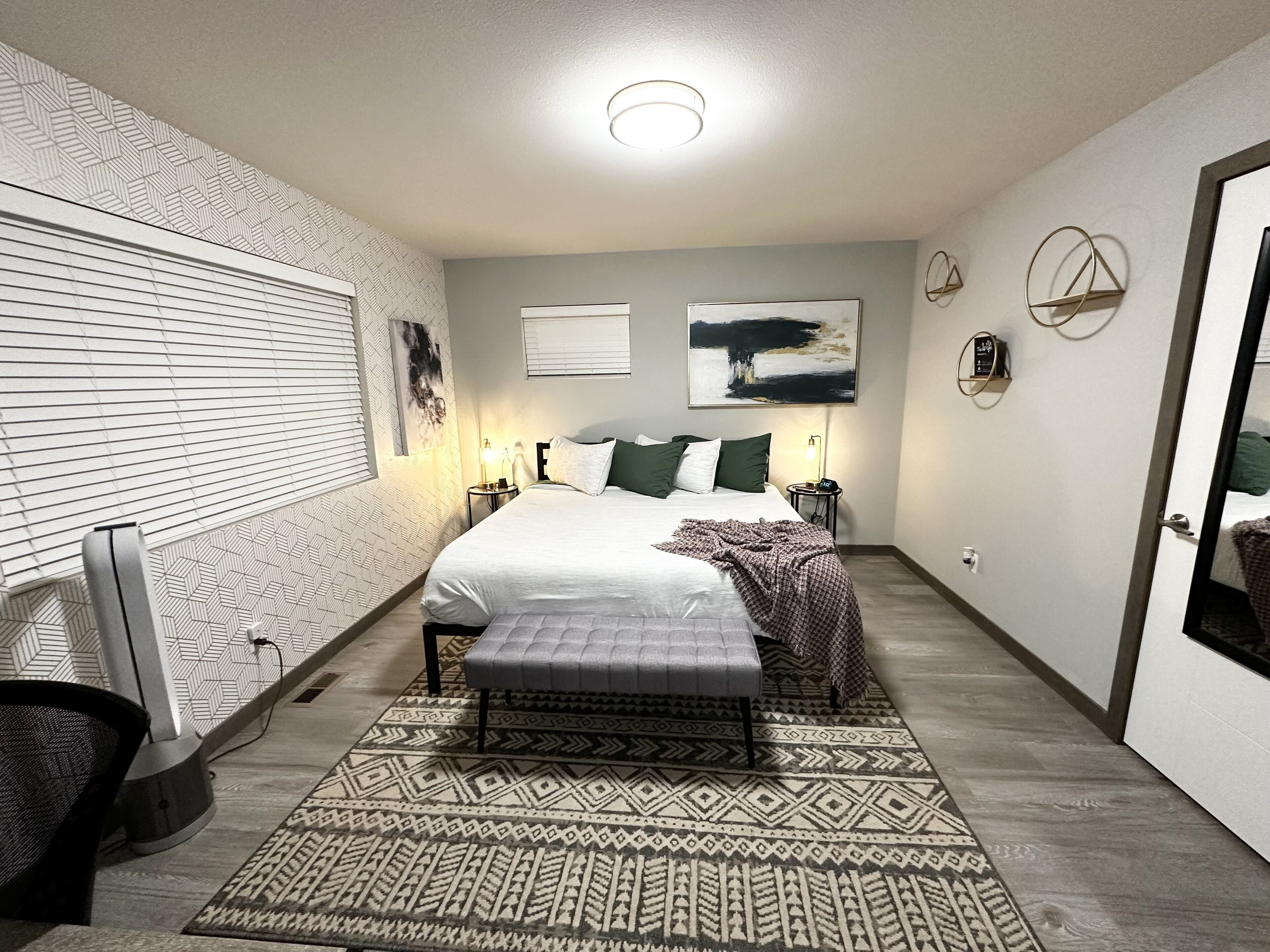 A modern bedroom with a large bed, a bedside table with a lamp, a wall-mounted TV, and a patterned rug.