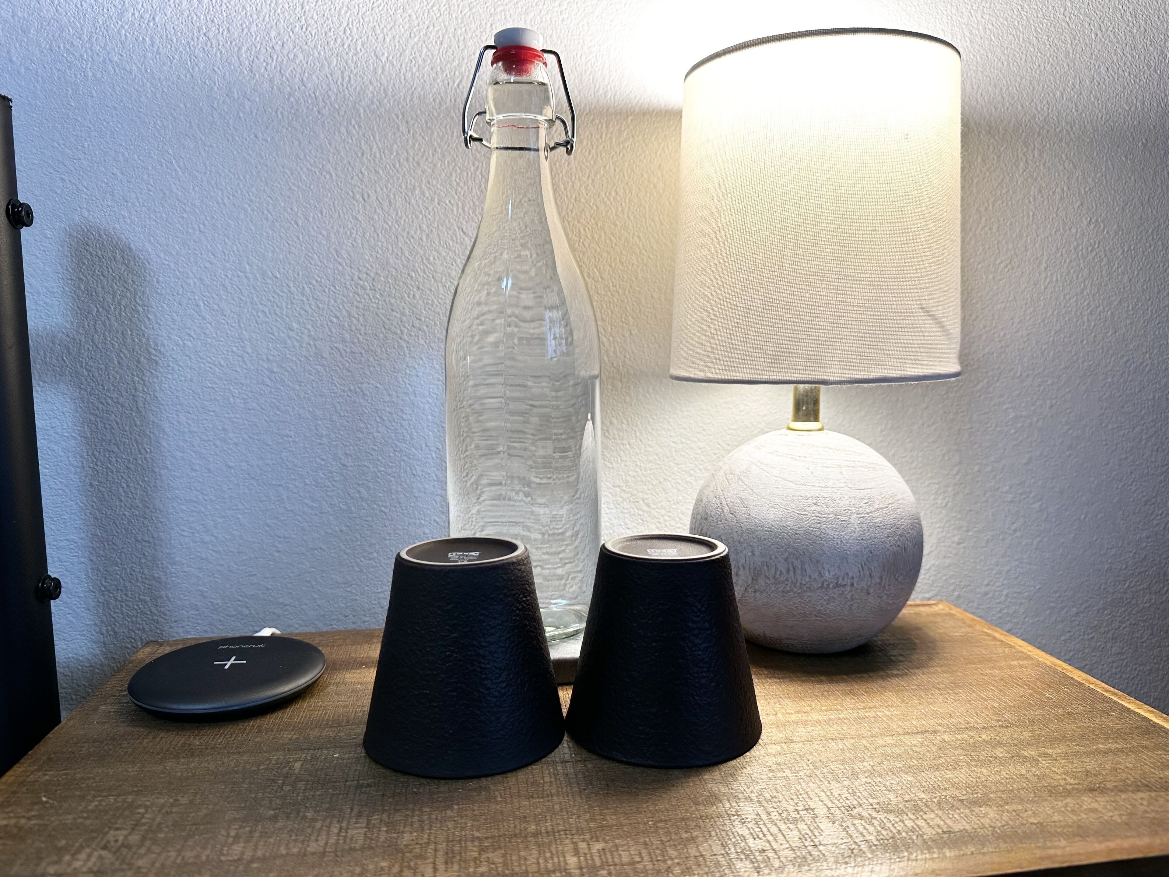 Een transparante glazen fles met een metalen sluiting, twee zwarte onderzetters en een lamp met een witte textuur, op een houten oppervlak voor een effen muur.