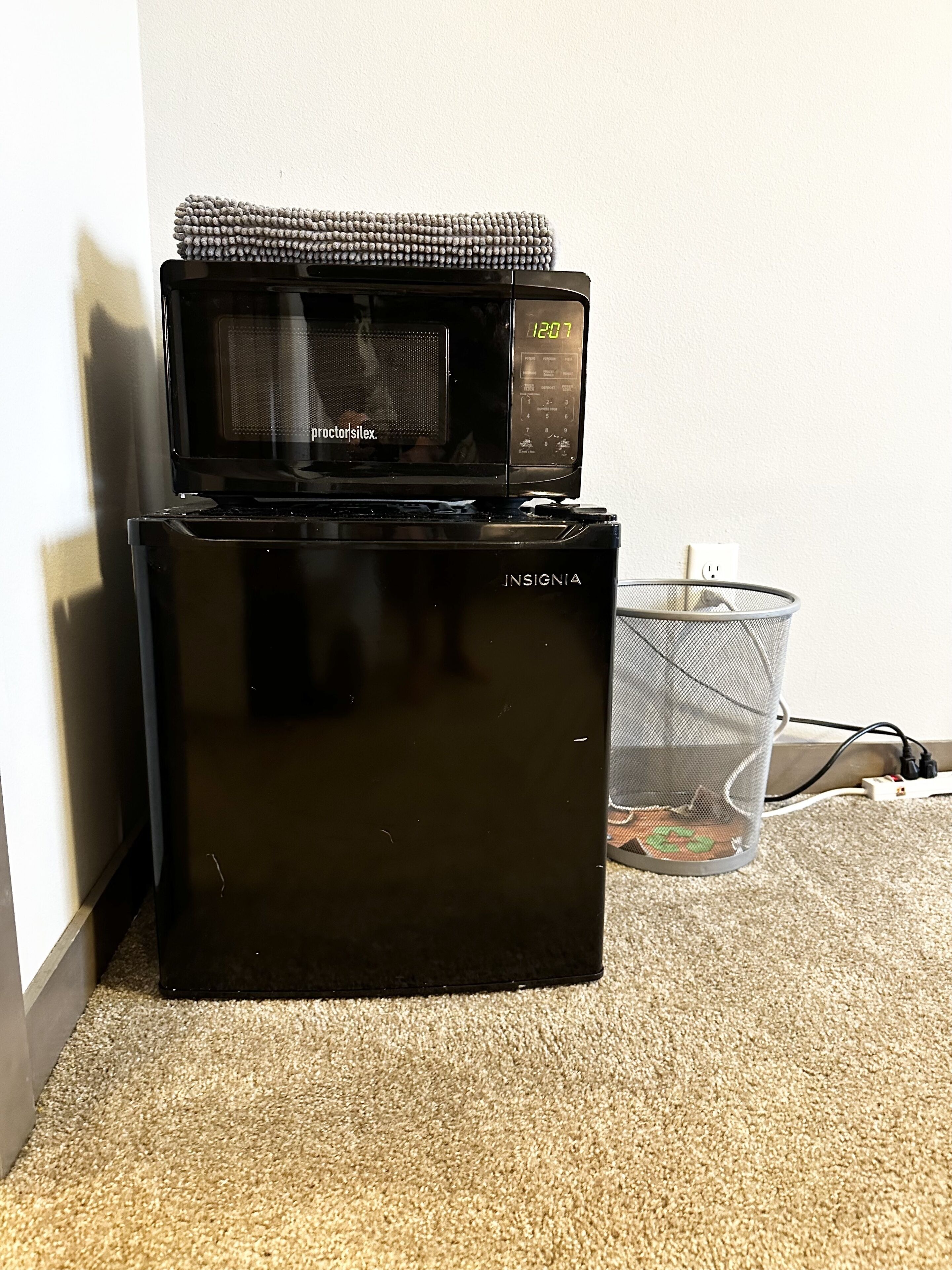 A black refrigerator with a microwave on top, a power outlet, and a mesh basket on the floor.