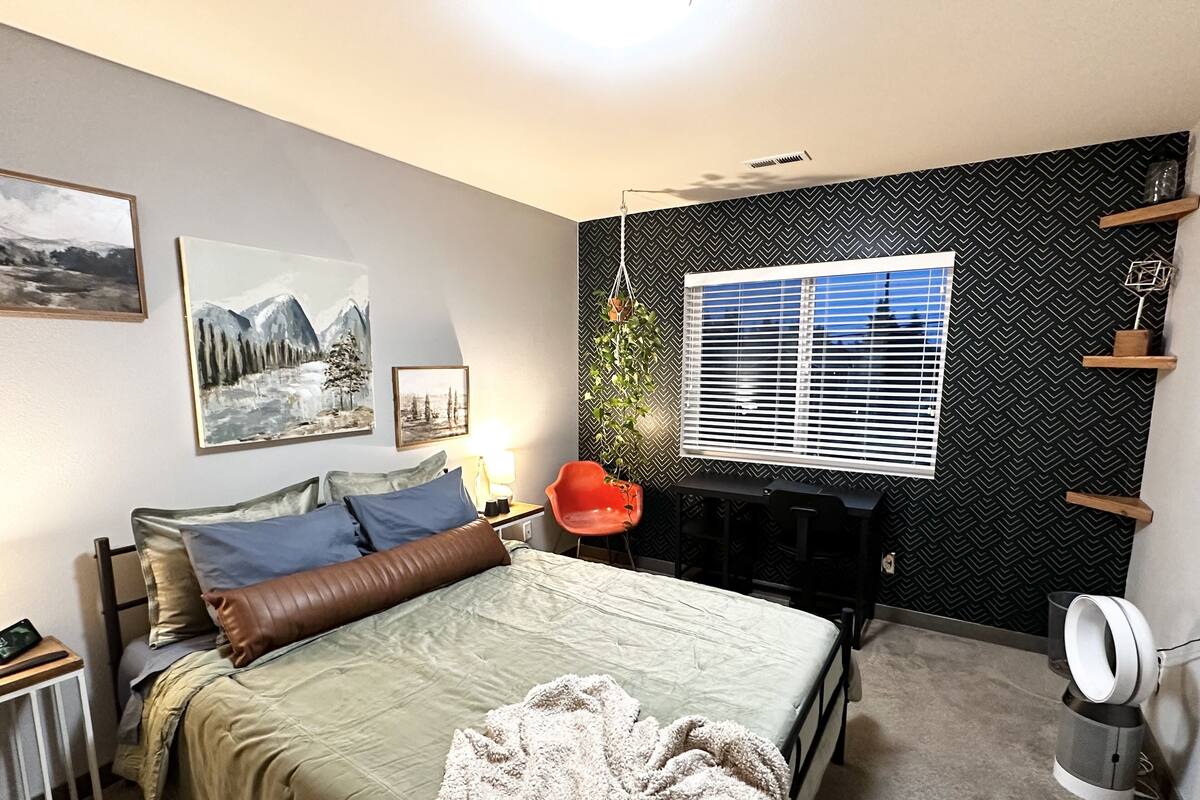 Ein Schlafzimmer mit Bett, Schreibtisch, Stuhl, einem Fenster mit Jalousien und Wandbildern.